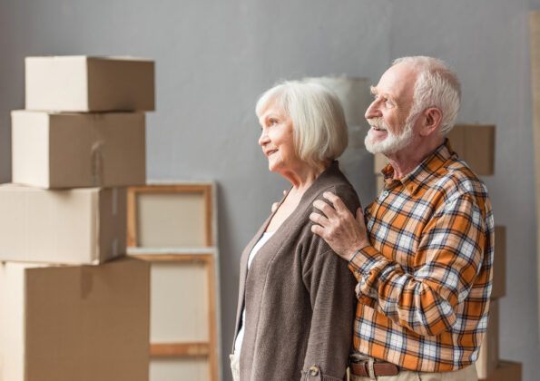 cheerful husband hugging wife shoulders in new house with cardboard boxes on background
